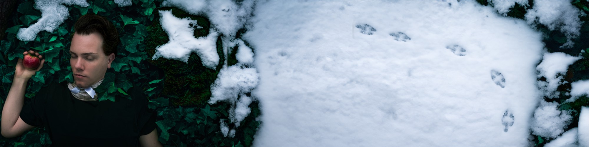 Mika Maria Schöberl liegt im Efeu und hält einen roten Apfel, daneben frischer Schnee mit Vogelspuren